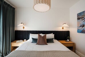 A hotel room with a large bed, two bedside lamps, a nightstand, a painting on the wall, and a window with curtains. - 2 Turns - Revelstoke Vacations (Revelstoke)
