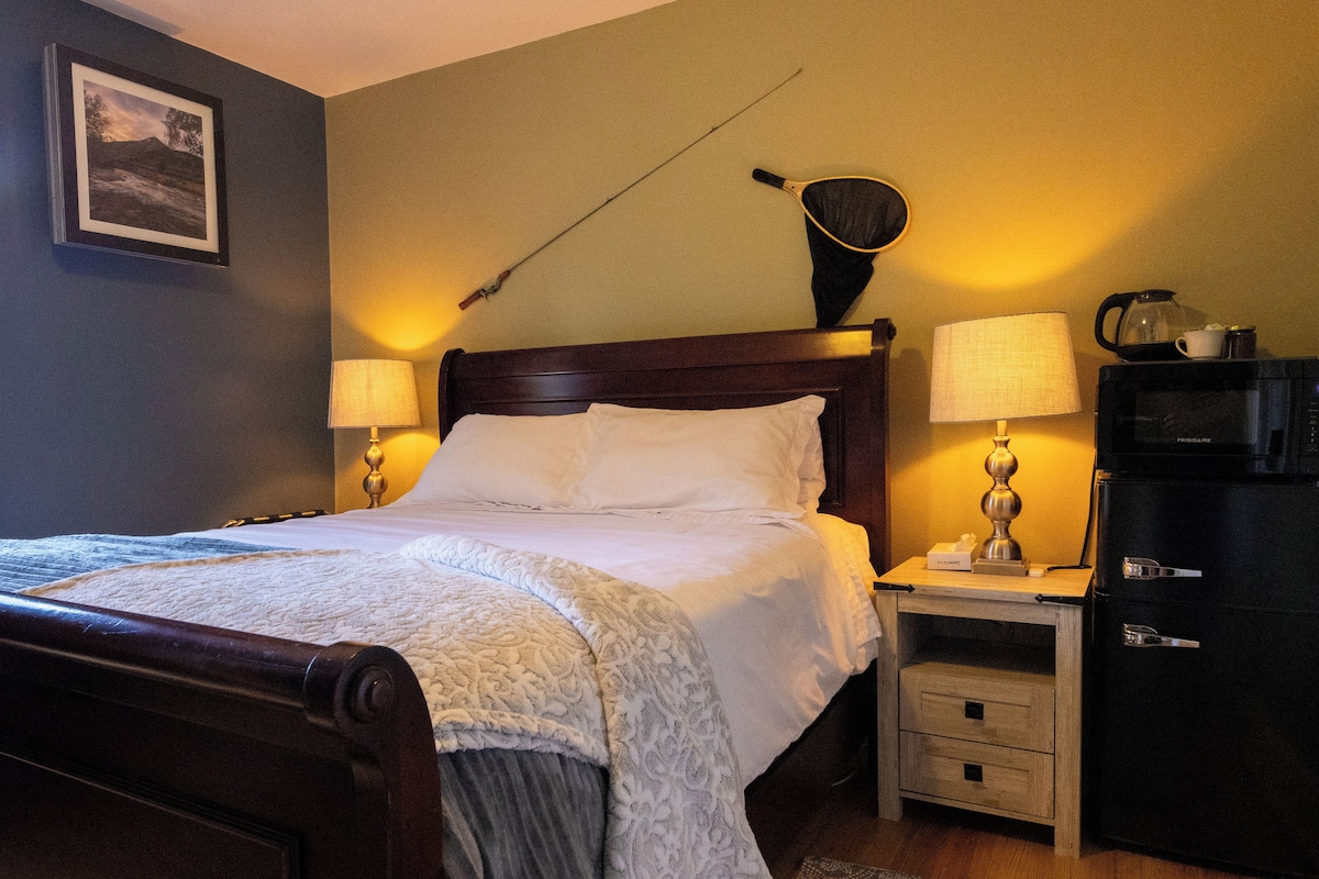 Una cama bien hecha con cabecera de madera, dos lámparas de noche, una mesita de noche con lámpara, un mueble pequeño y un cuadro en la pared.