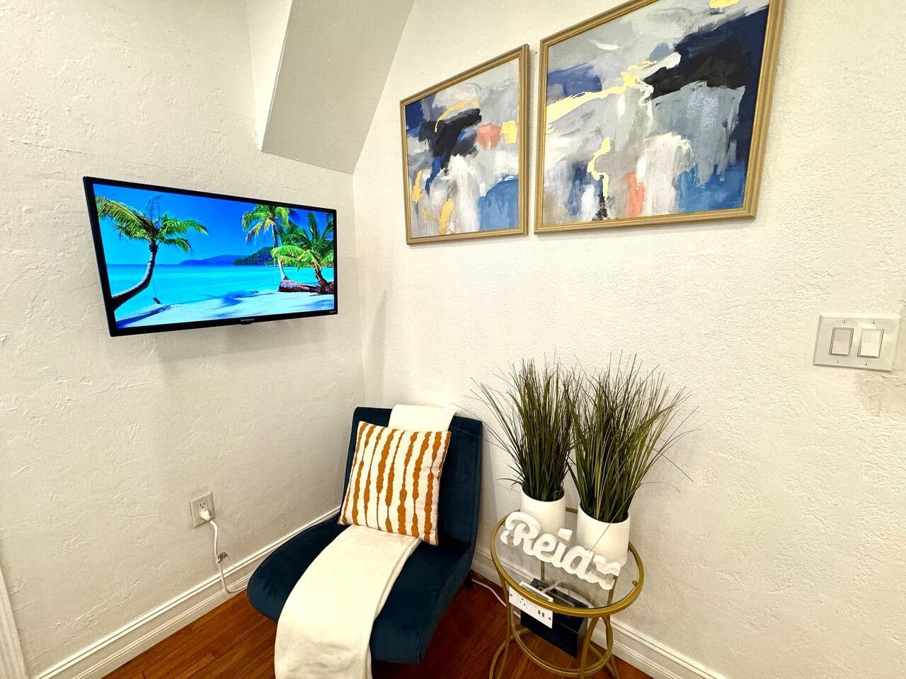 A living room with a flat-screen TV, a blue sofa with patterned cushions, and two abstract paintings on the wall.