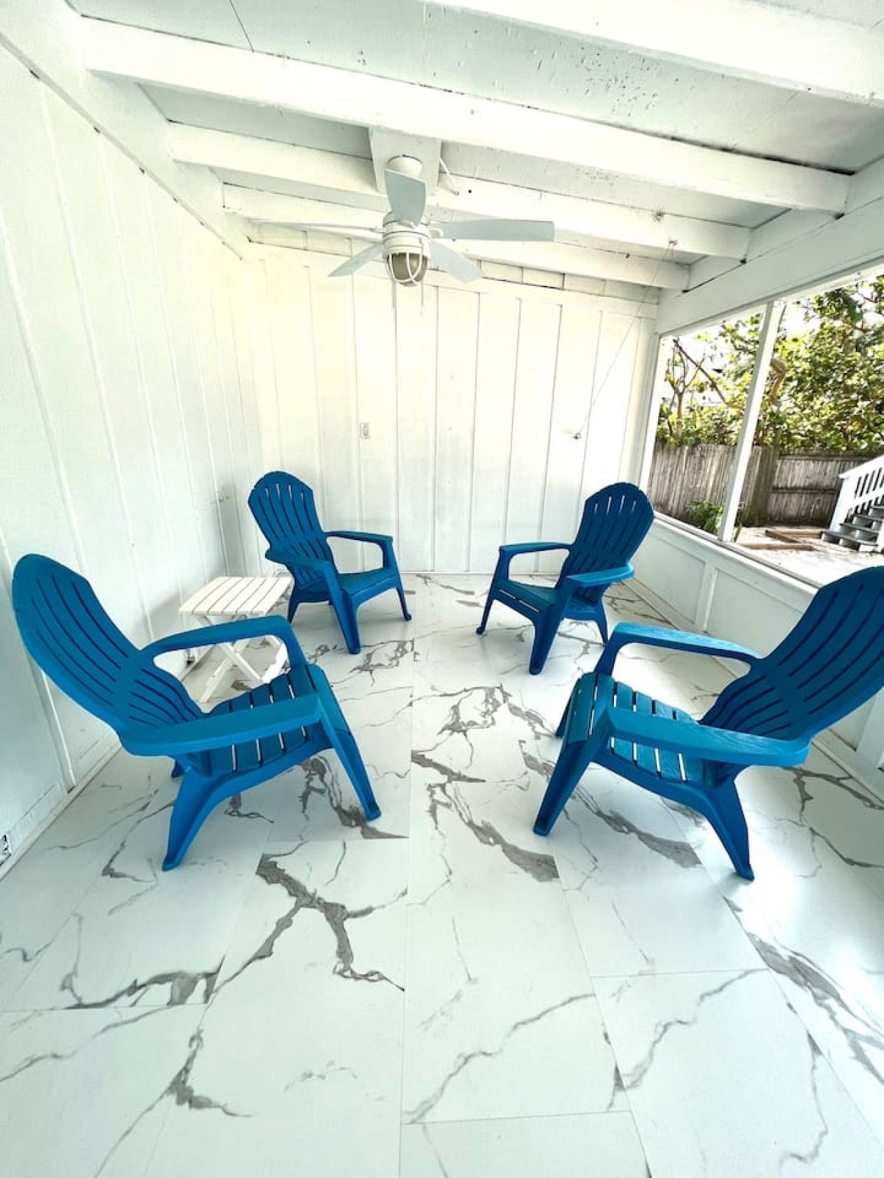 Una veranda riparata con quattro sedie blu e un tavolo, pareti bianche e una ventola a soffitto.