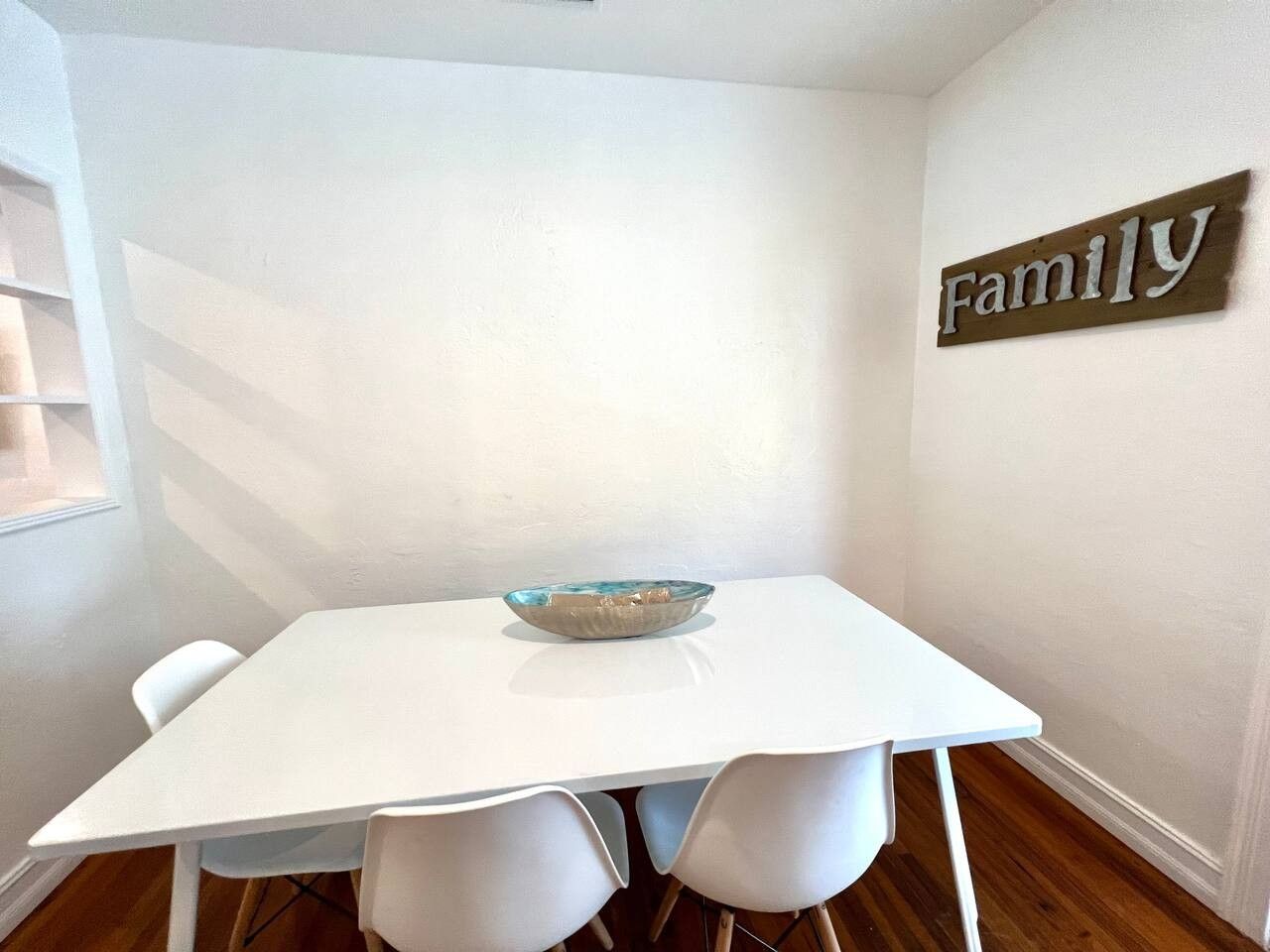Une table à manger blanche, avec un saladier au centre, entourée de chaises blanches, le tout placé devant un mur blanc sur lequel est accroché un panneau indiquant « Famille ».