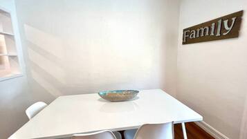Une table à manger blanche, avec un saladier au centre, entourée de chaises blanches, le tout placé devant un mur blanc sur lequel est accroché un panneau indiquant « Famille ».