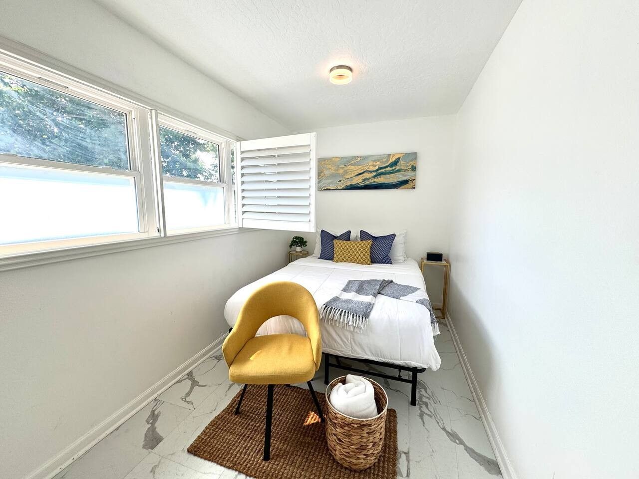 Une chambre à coucher comprenant un lit, une chaise, une table de chevet, une œuvre d’art accrochée au mur et une fenêtre avec des stores.