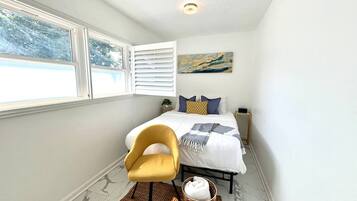 Une chambre à coucher comprenant un lit, une chaise, une table de chevet, une œuvre d’art accrochée au mur et une fenêtre avec des stores.