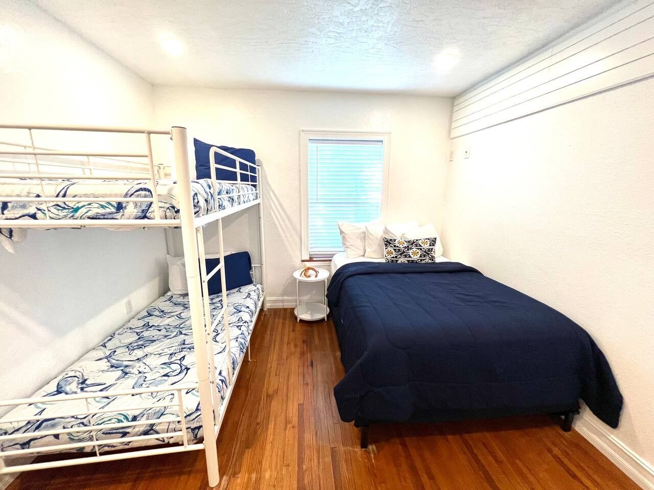 A room with bunk beds, a single bed, a small bedside table, and a window with blinds.