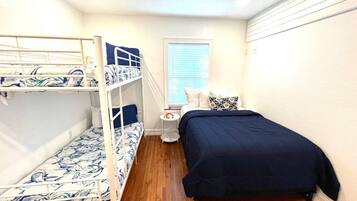 A room with bunk beds, a single bed, a small bedside table, and a window with blinds.
