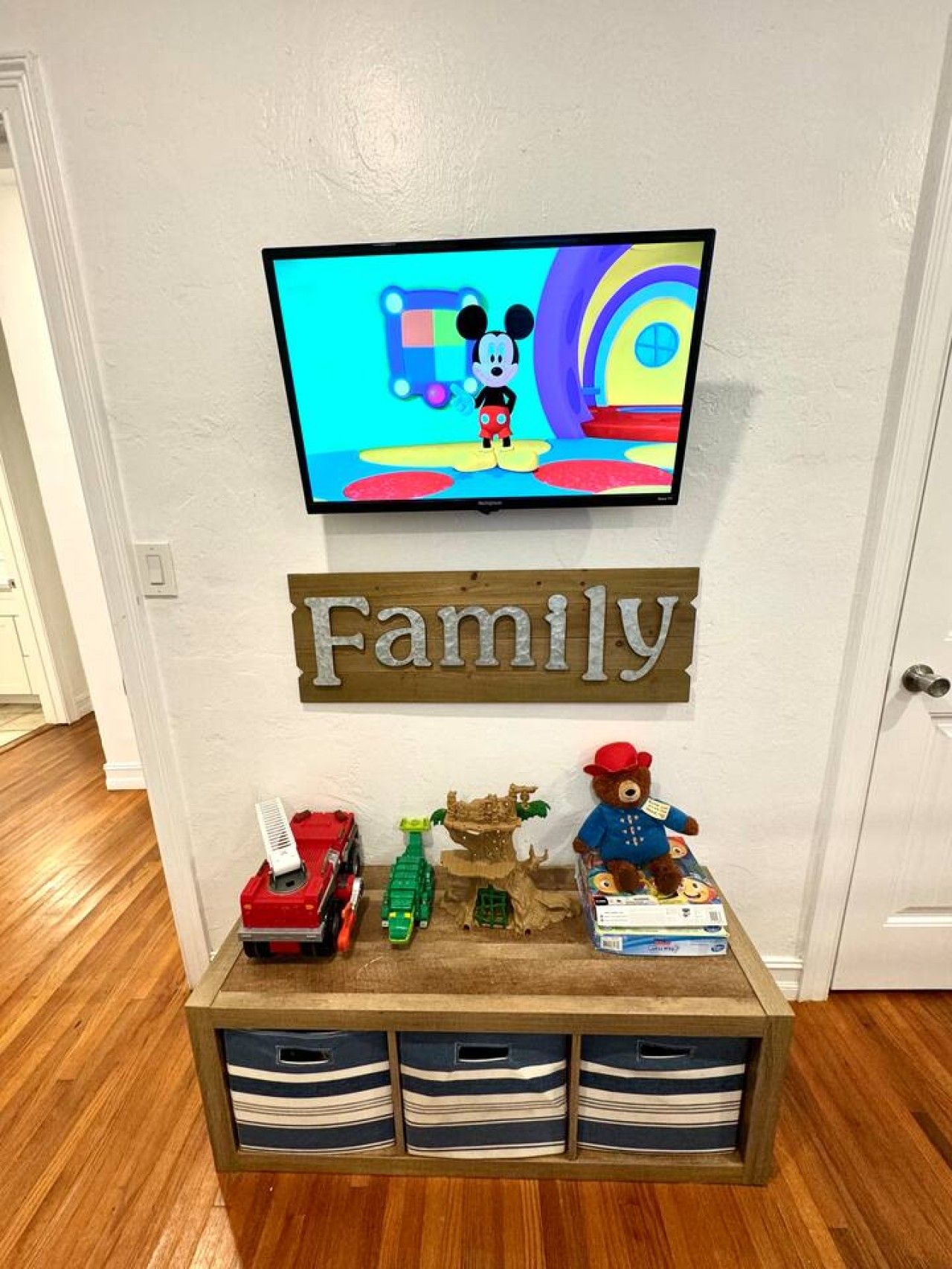 A wall-mounted television displaying a cartoon character, a wooden sign with the word 'Family,' and a shelf with toys and storage bins.