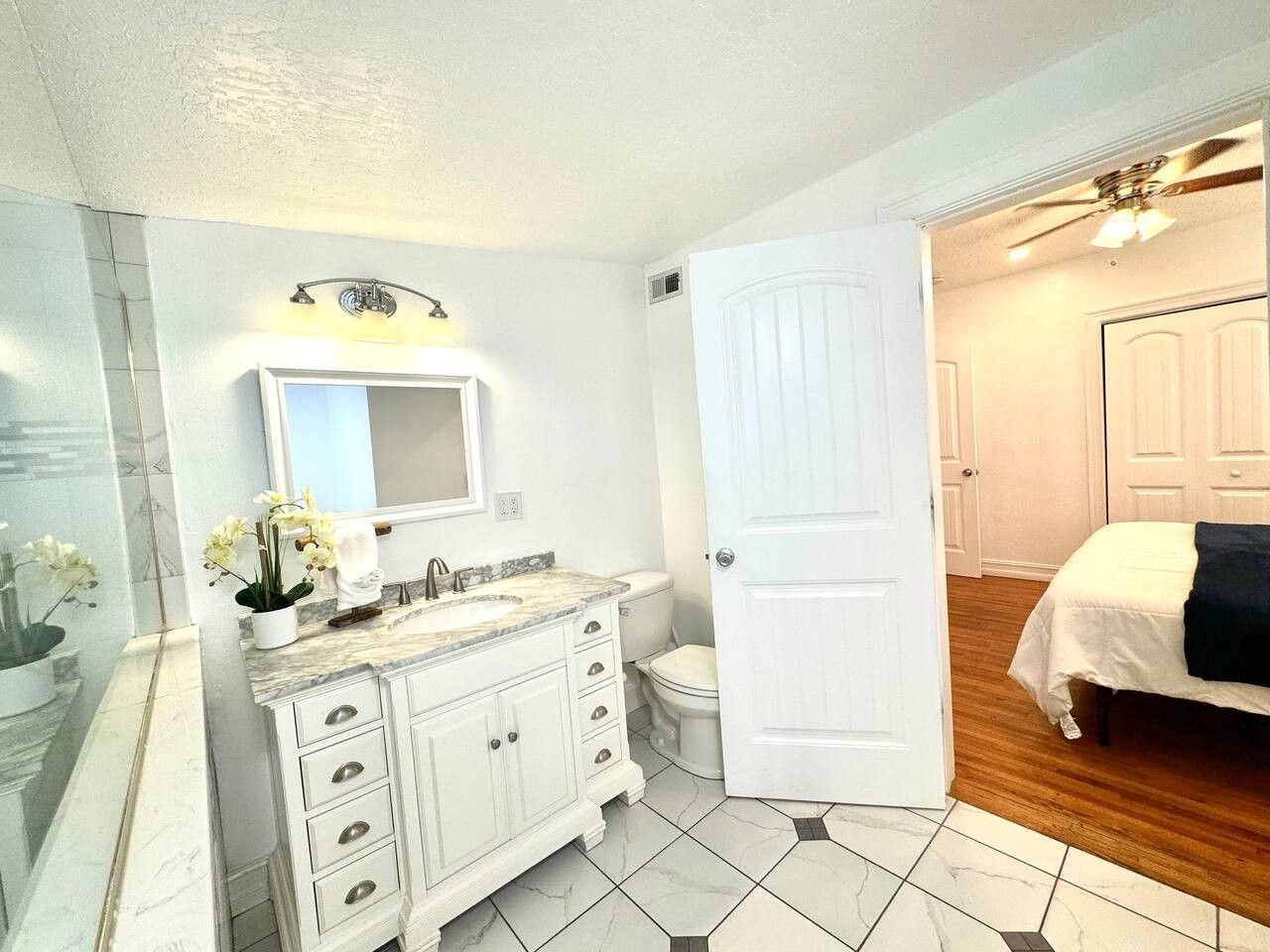 Une salle de bain avec un meuble-lavabo blanc, un évier, un miroir et des toilettes. Une porte donne sur une chambre à coucher où se trouve un lit.