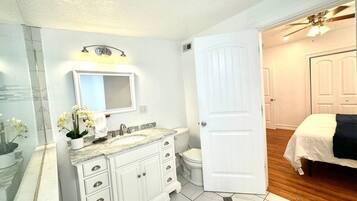 Une salle de bain avec un meuble-lavabo blanc, un évier, un miroir et des toilettes. Une porte donne sur une chambre à coucher où se trouve un lit.