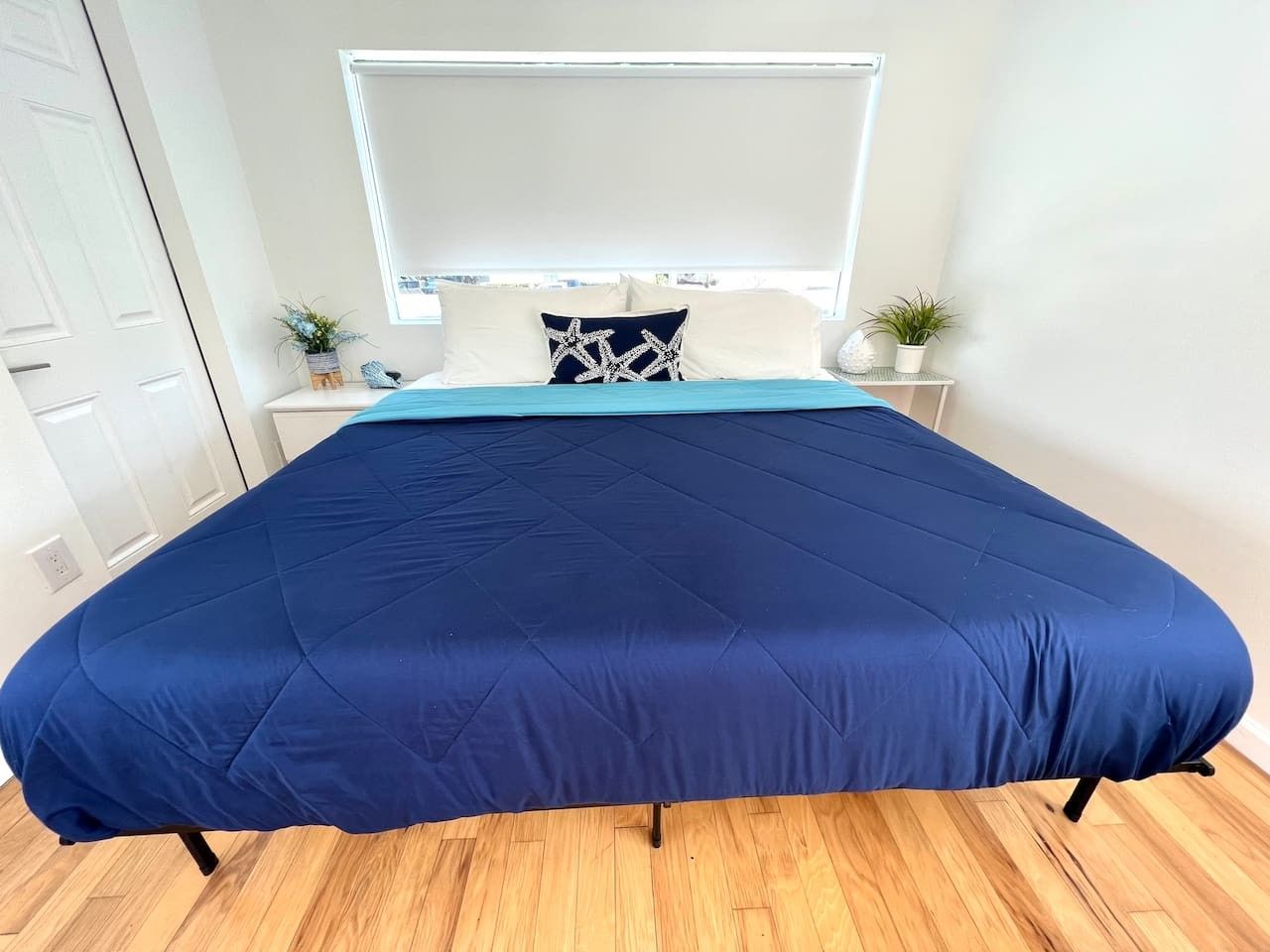 Una cama con colcha azul, cabecera blanca y una mesita de noche con una planta.