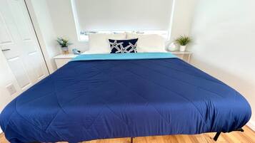 Una cama con colcha azul, cabecera blanca y una mesita de noche con una planta.