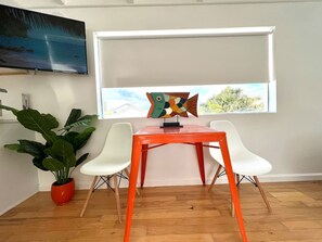 Een kamer met een houten vloer, een rode tafel, twee witte stoelen, een potplant, een kleurrijke vissenbeeld en een aan de muur bevestigde televisie.