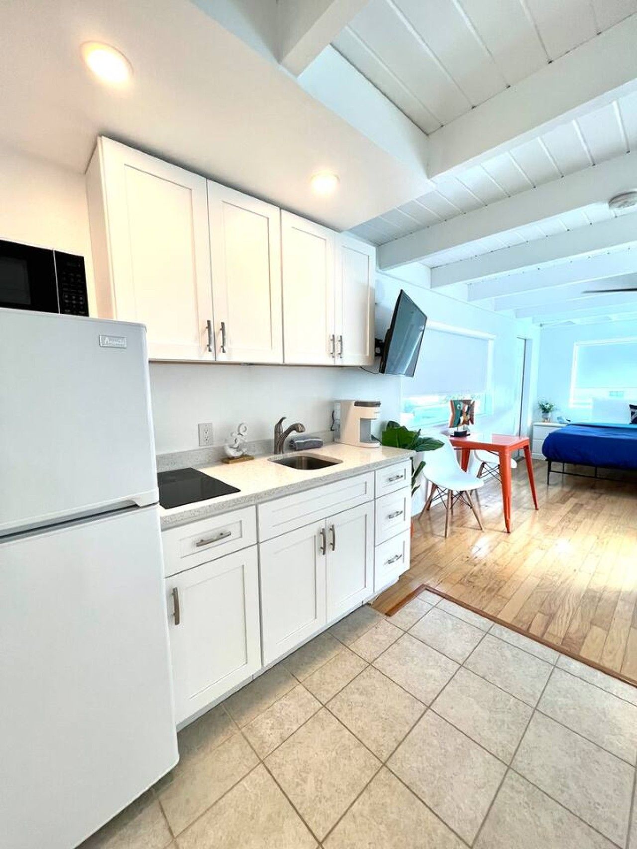 Una cocina moderna con armarios blancos, un microondas integrado y un lavamanos. Hay una zona de comedor con una mesa y sillas, y un televisor montado en la pared.