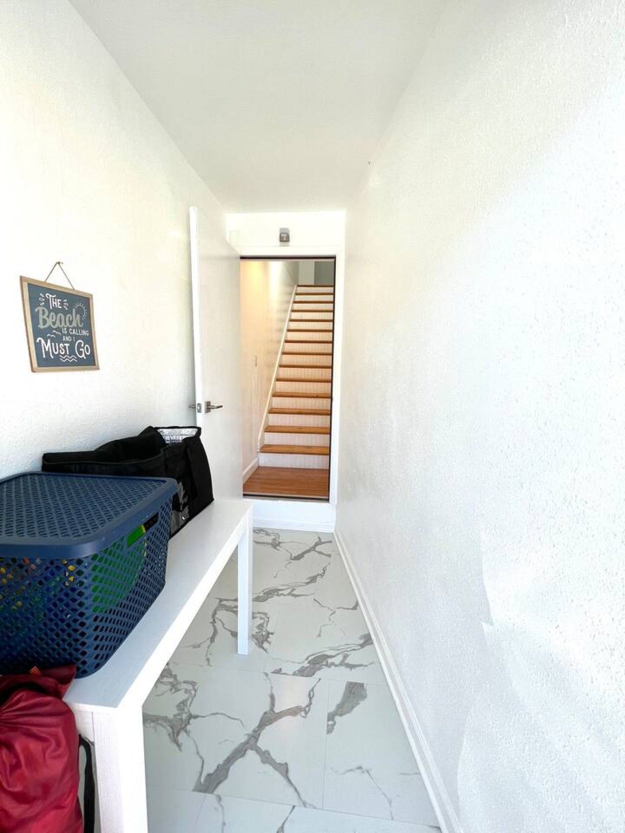 Un pasillo con una escalera que sube a un piso superior, un letrero en la pared, una cesta y un bolso en un estante.