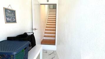 Un pasillo con una escalera que sube a un piso superior, un letrero en la pared, una cesta y un bolso en un estante.