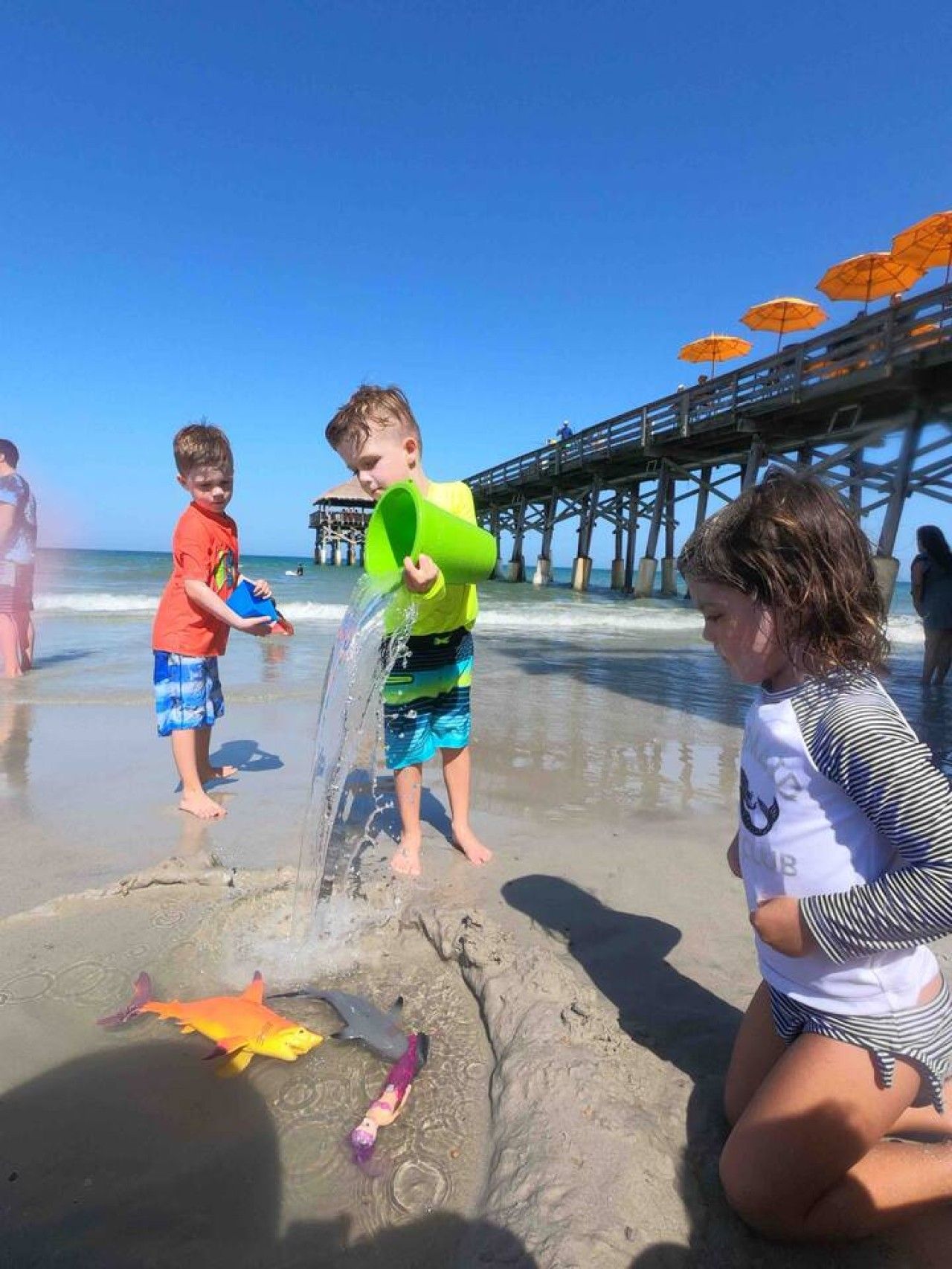 Niños jugando en una playa arenosa con un muelle al fondo.