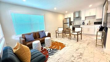 A modern living room with a sofa, coffee table, and patterned rug. A kitchen with white cabinets and a microwave is visible in the background.