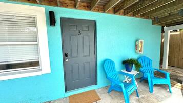 Pared exterior turquesa con una puerta gris oscuro, una ventana con persianas y dos sillas de plástico azul con una planta en maceta.