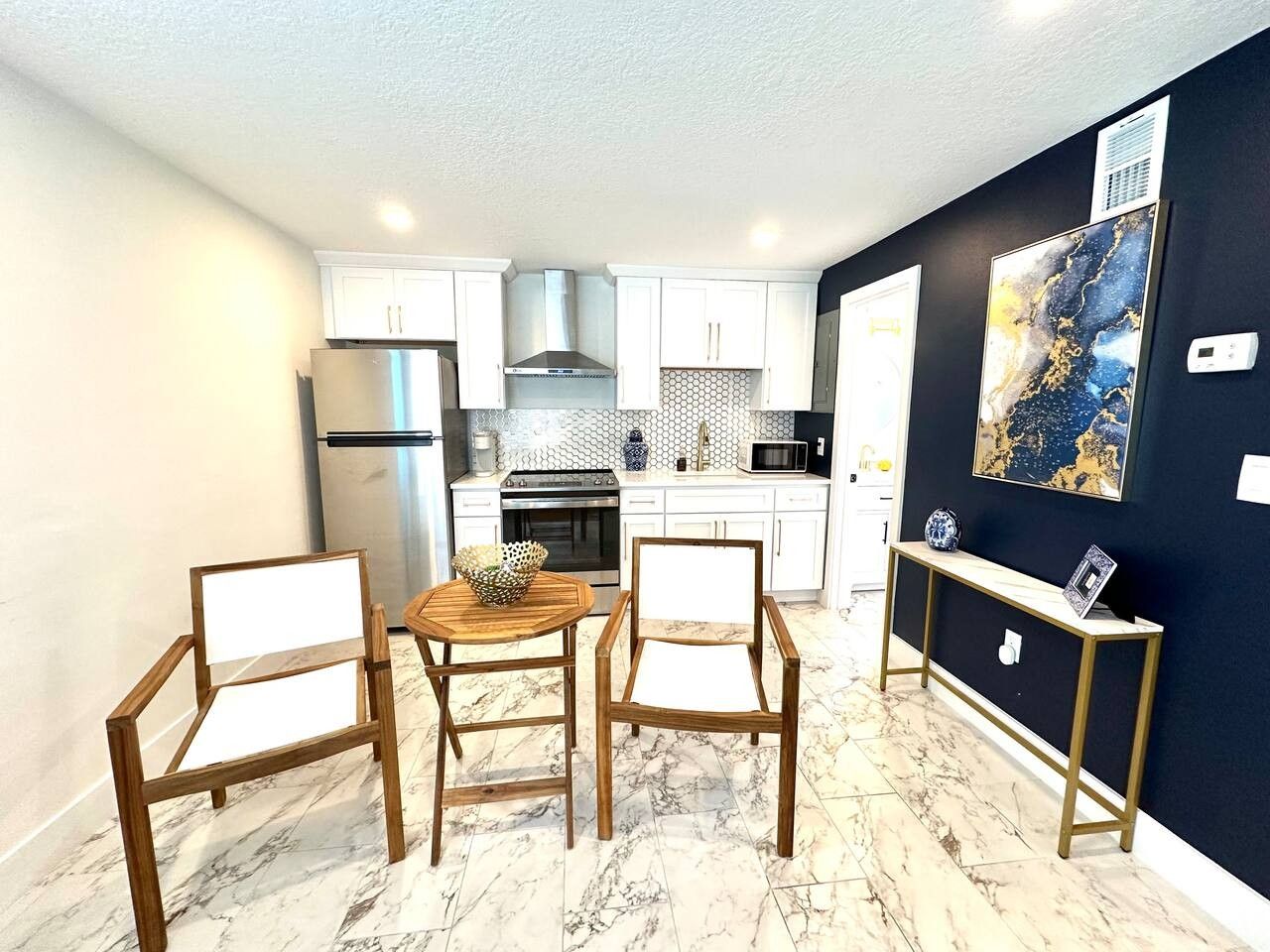 Cocina compacta con armarios blancos, refrigerador de acero inoxidable, microondas y estufa. Hay un comedor con mesa y sillas de madera.