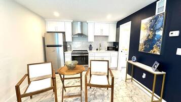 Cocina compacta con armarios blancos, refrigerador de acero inoxidable, microondas y estufa. Hay un comedor con mesa y sillas de madera.