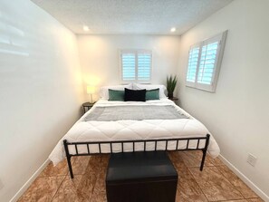 Ein Schlafzimmer mit Bett, Nachttisch, Lampe, Fenster und Pflanze.