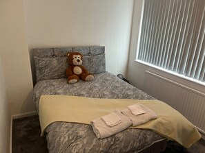 Ein Bett mit einer grauen, gemusterten Tagesdecke, einem braunen Teddybären und einer gelben Decke mit gefalteten Handtüchern.