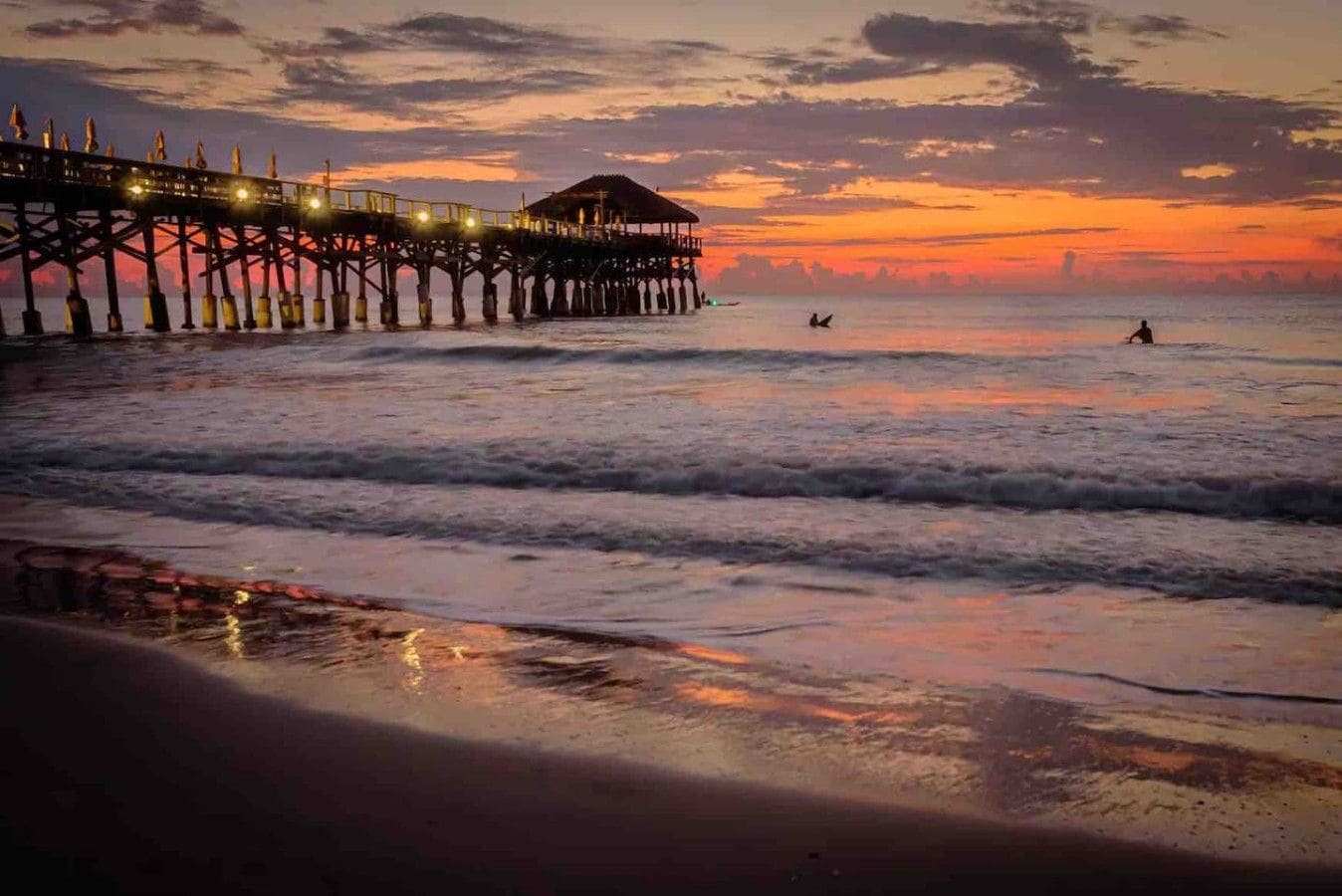 A pier extending into the water with a structure at the end, waves gently lapping at the shore, and a clear sky above.