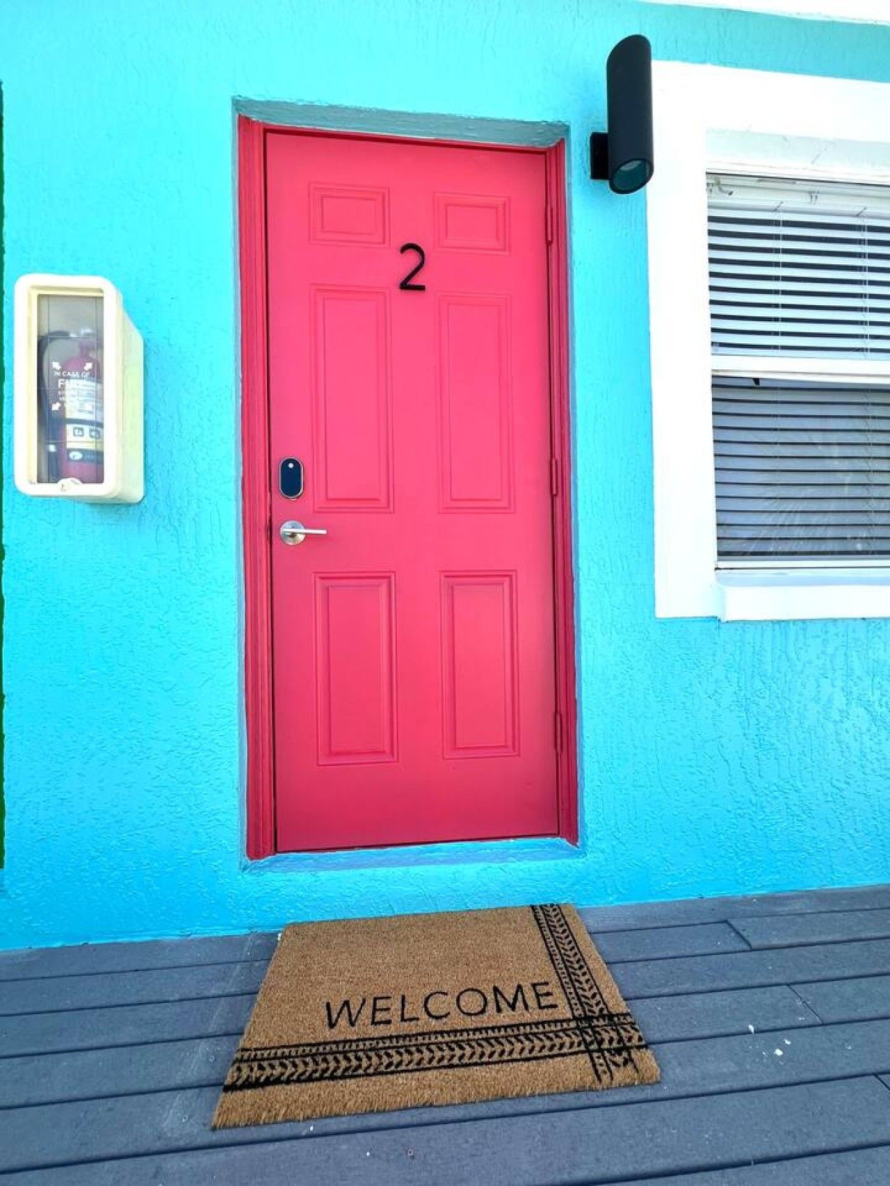 Una puerta roja con el número 2, un buzón y un felpudo sobre una terraza de madera.