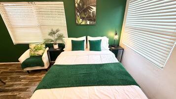 Un dormitorio con cama, una silla, mesita de noche, lámpara y ventana con persianas.