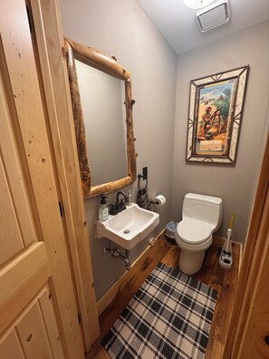 A bathroom with a wooden door, a white sink, a mirror, a toilet, and a framed picture on the wall. - Secluded 5-BR Wooded Cabin Retreat with Panoramic Views in Vanderbilt (Vanderbilt)