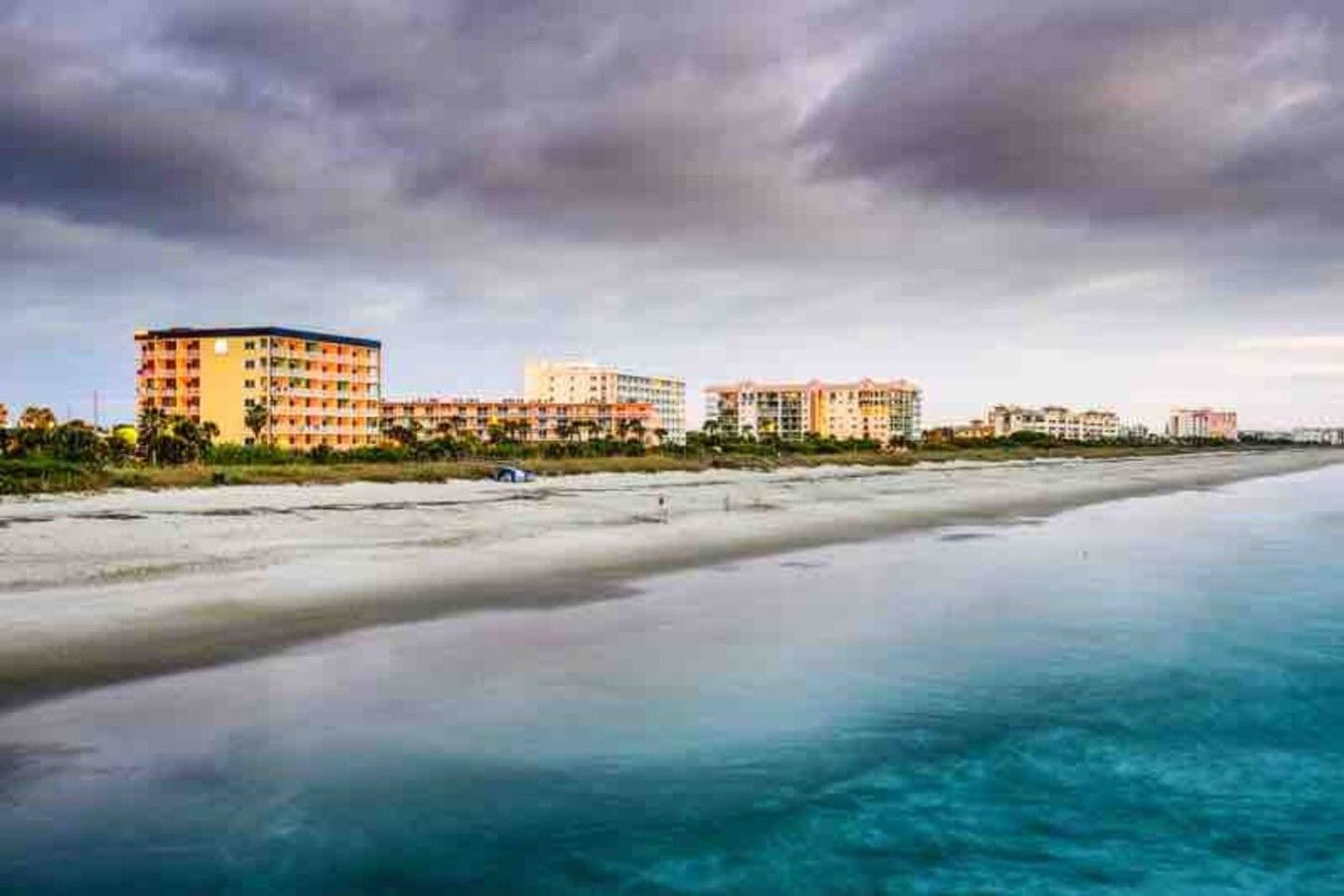 Un hotel frente a la playa con varias construcciones, una playa de arena y aguas tranquilas.