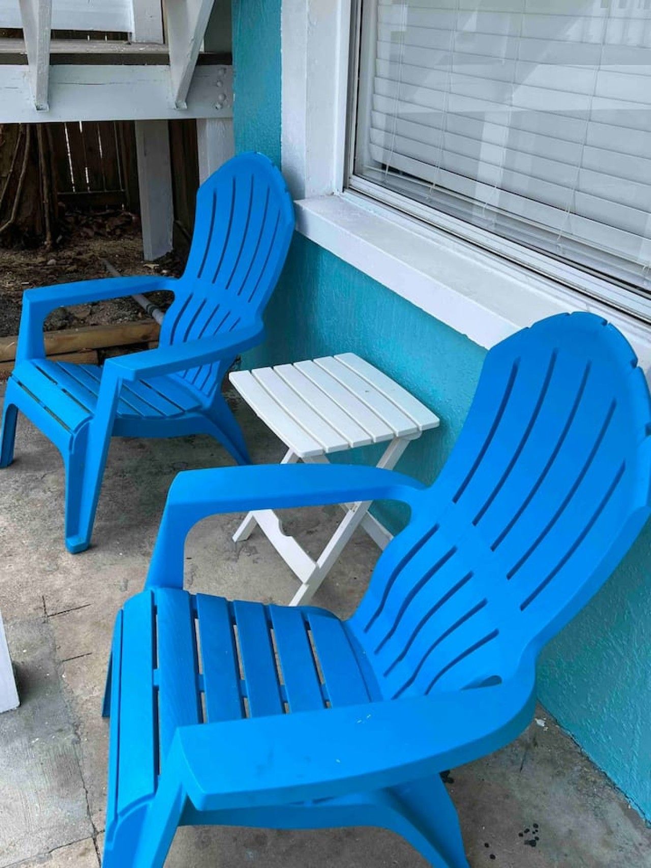 Dos sillas Adirondack azules y una mesita blanca frente a un edificio con una ventana.