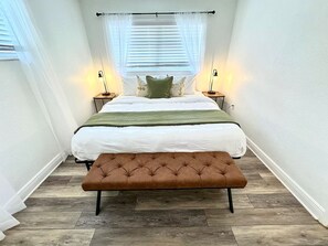 A bedroom with a bed, bench, bedside lamps, and a window with blinds. - 1 bdrm:beach across st, Port  8 mi, Ron Jon’s 4 mi (Cocoa Beach)