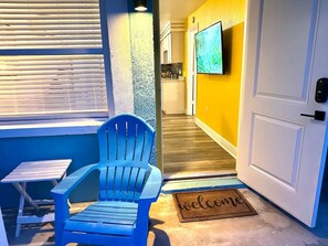 A blue chair outside a door with a 'welcome' mat, a small table, and a TV on the wall. - 1 bdrm:beach across st, Port  8 mi, Ron Jon’s 4 mi (Cocoa Beach)