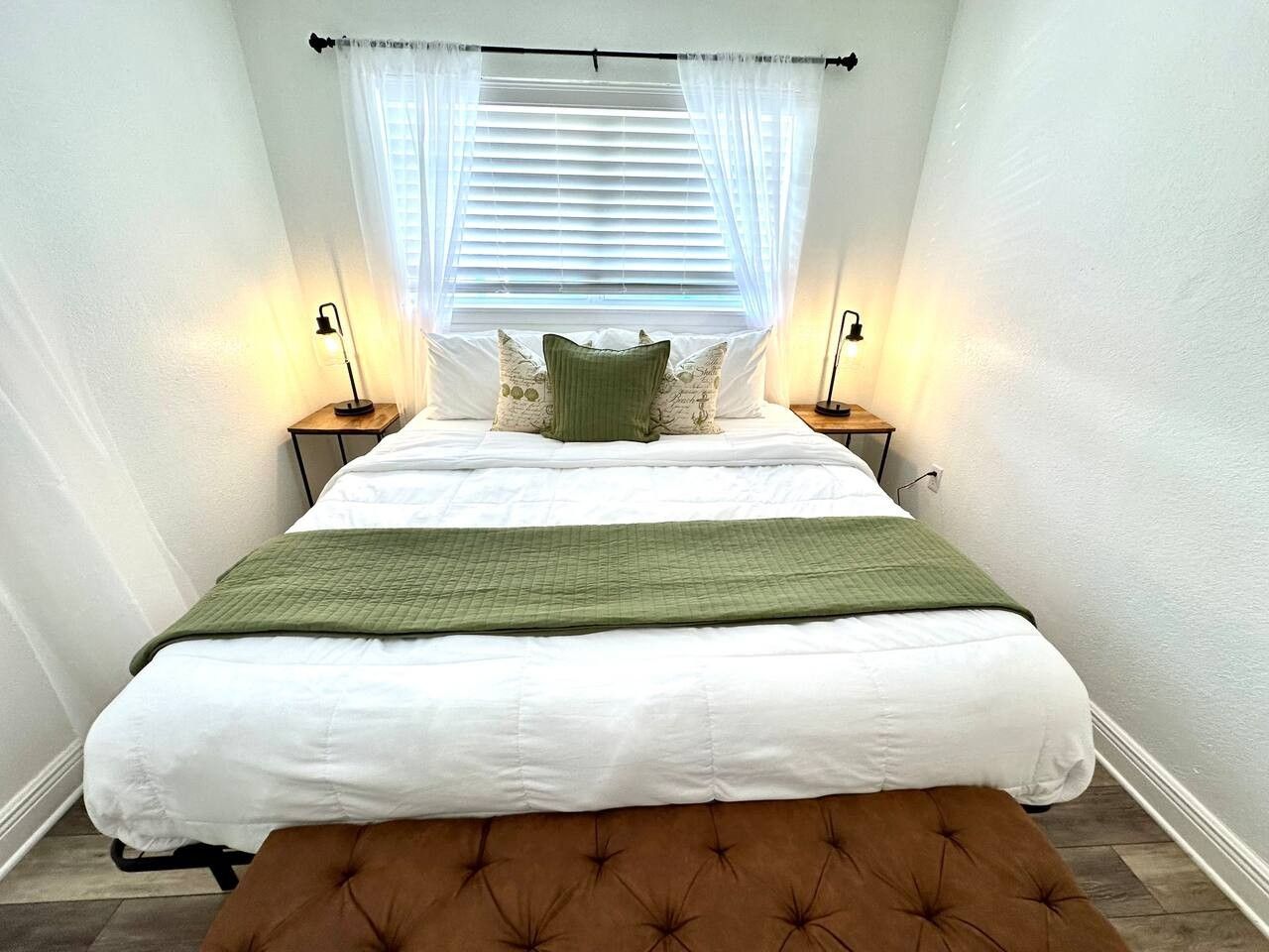 Un dormitorio con cama, dos mesitas de noche, un banco, una ventana con persianas y dos lámparas de pared.