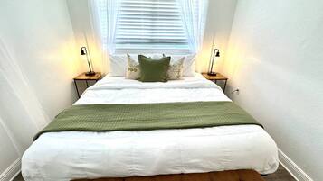 Un dormitorio con cama, dos mesitas de noche, un banco, una ventana con persianas y dos lámparas de pared.