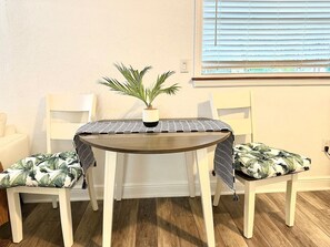A round dining table with a woven table runner, two chairs with tropical leaf-patterned cushions, and a potted plant on the table. - 1 bdrm:beach across st, Port  8 mi, Ron Jon’s 4 mi (Cocoa Beach)
