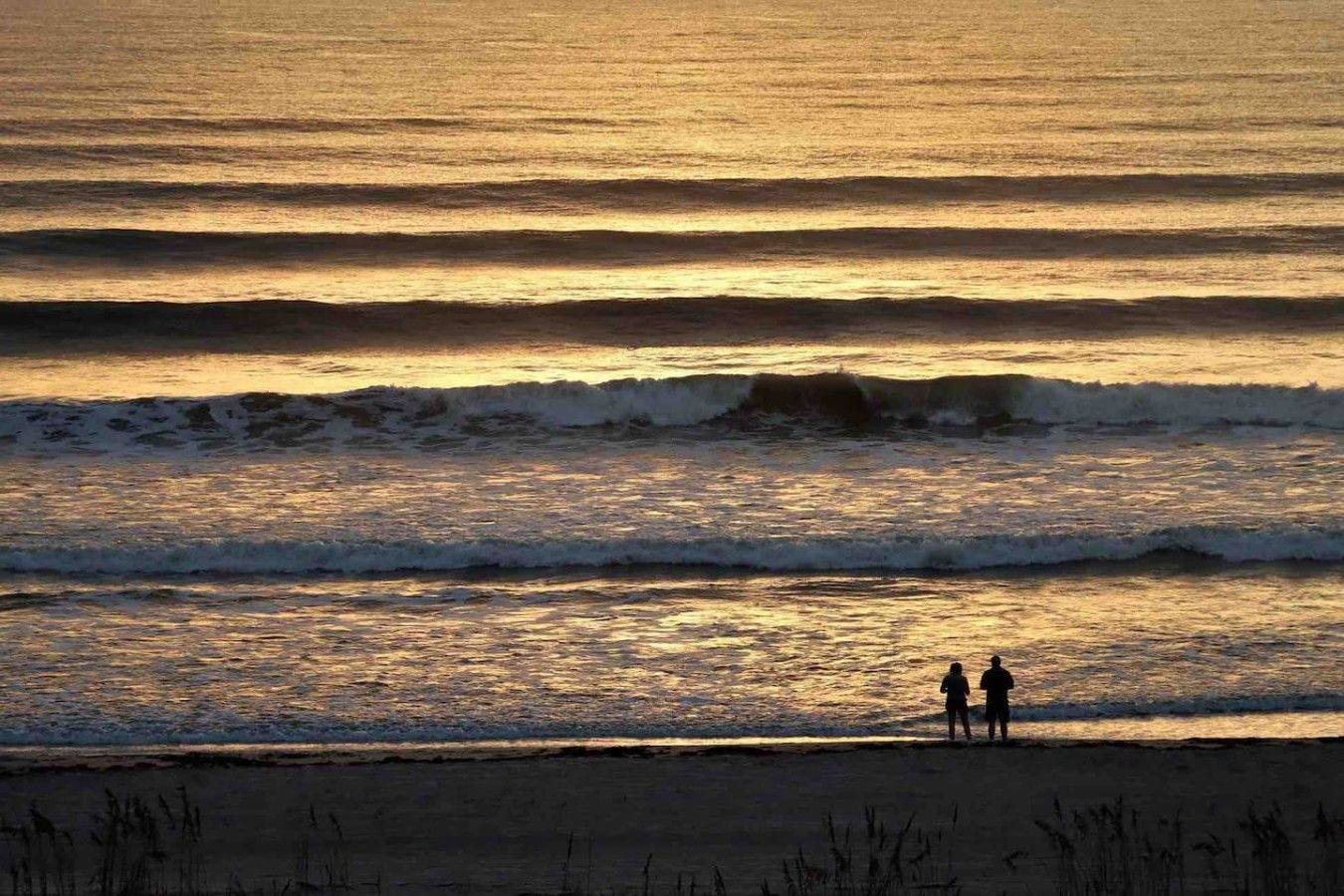 A beach scene with waves and two people standing on the shore.