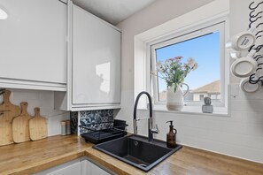A modern kitchen with a black sink, wooden cutting boards, a window with a view, and a cactus on the windowsill. - Premium 2 Bedroom Flat • ExCeL • Free Parking (Greater London)