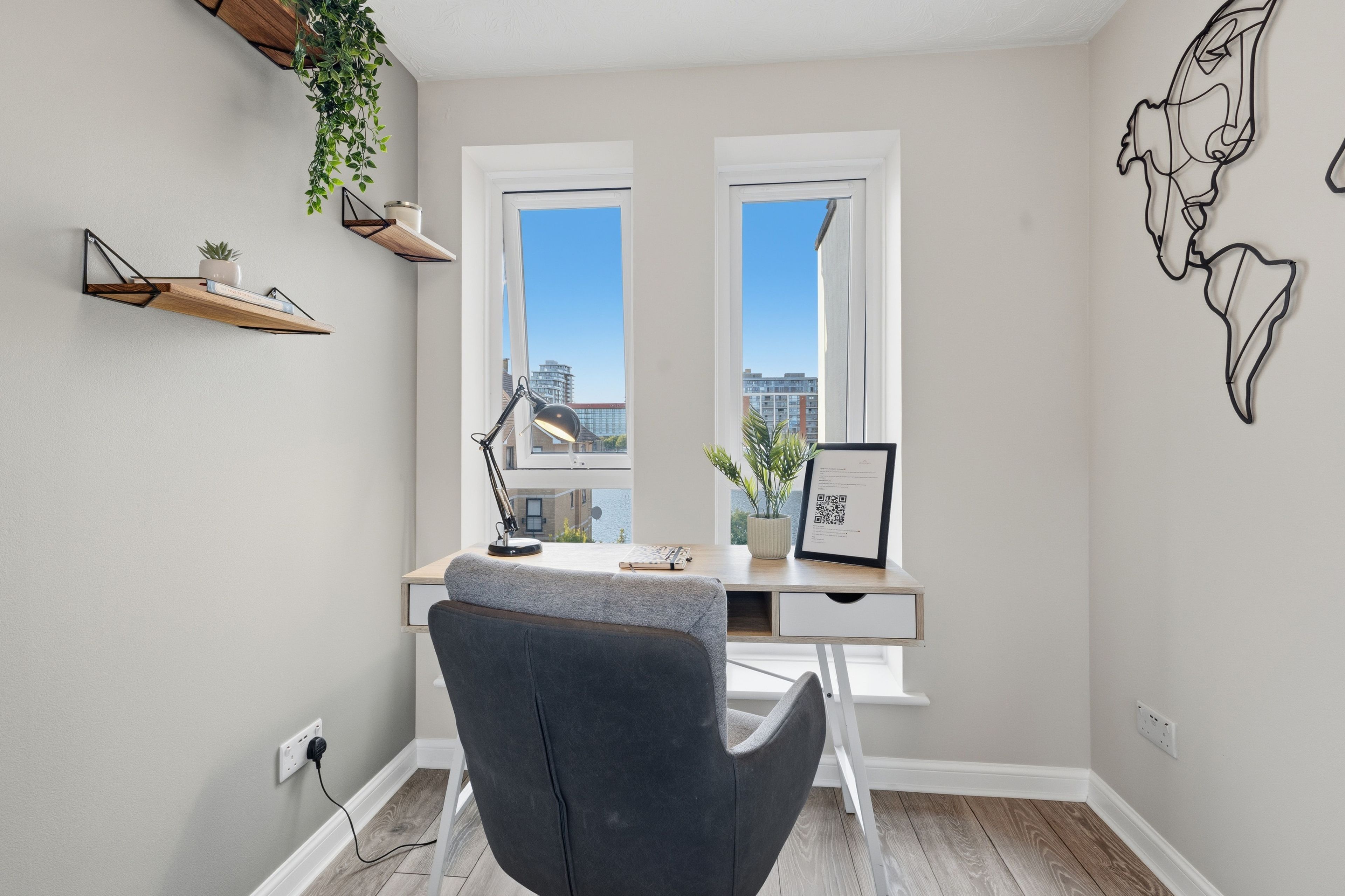 A modern office space with a desk, chair, two windows, a hanging plant, and a wall art piece.