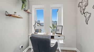 A modern office space with a desk, chair, two windows, a hanging plant, and a wall art piece.