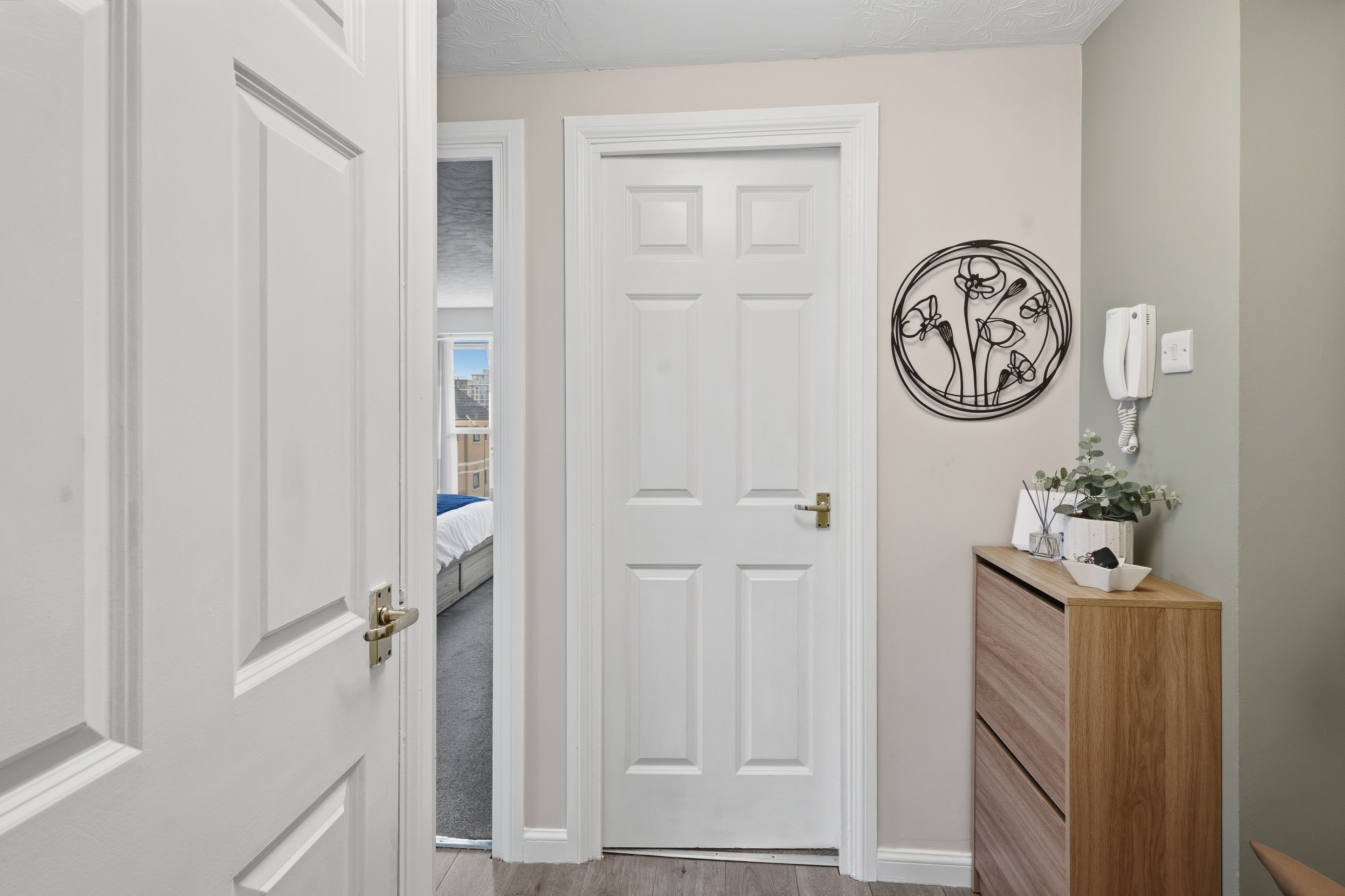 A hallway with a white door, a wooden dresser, a wall-mounted phone, and a circular artwork.