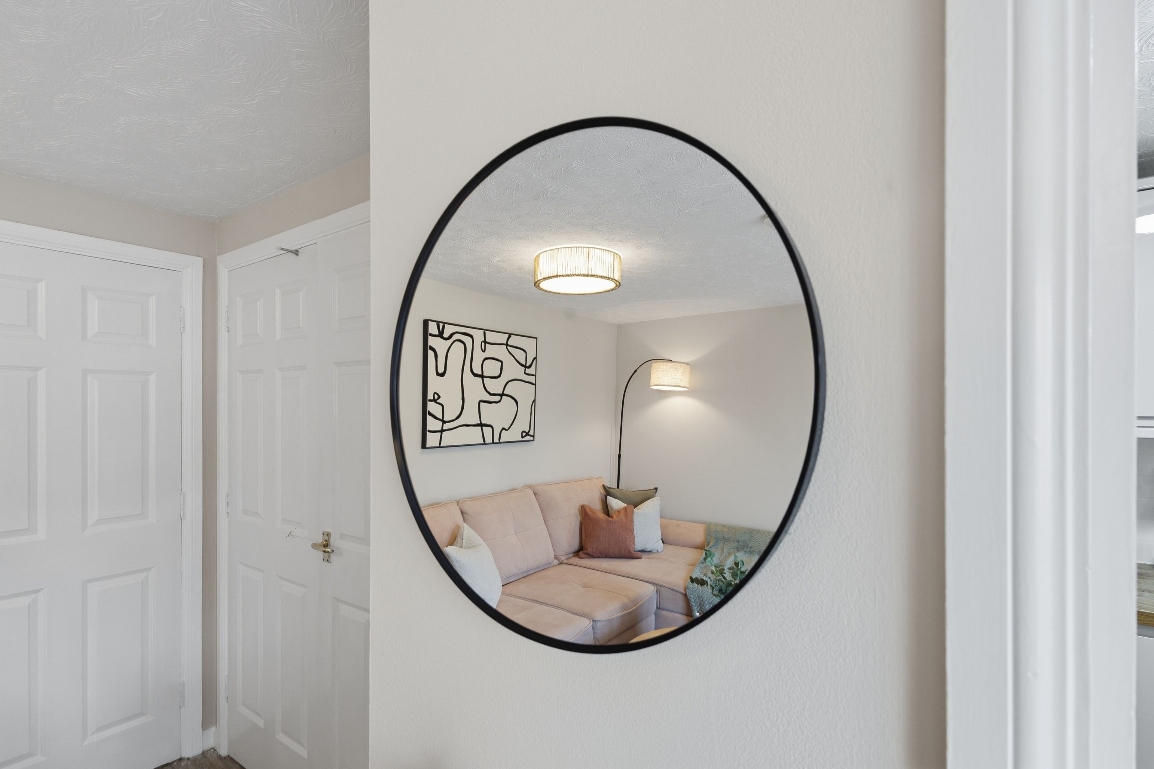 A hallway with a white door, a round mirror on the wall, a light fixture, and a piece of abstract art.