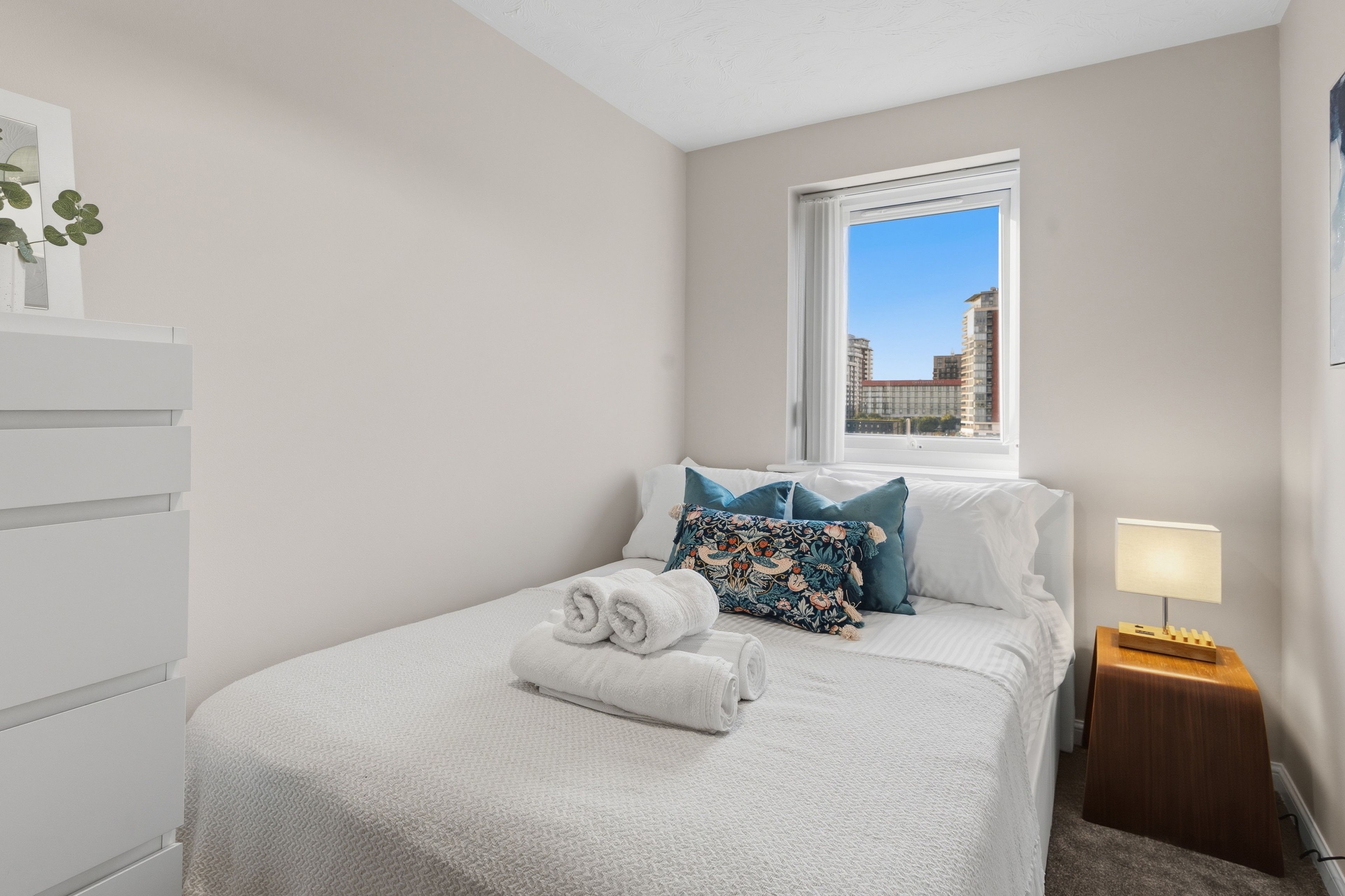 A bedroom with a bed, a window, a nightstand, and a wall-mounted shelf with a plant.