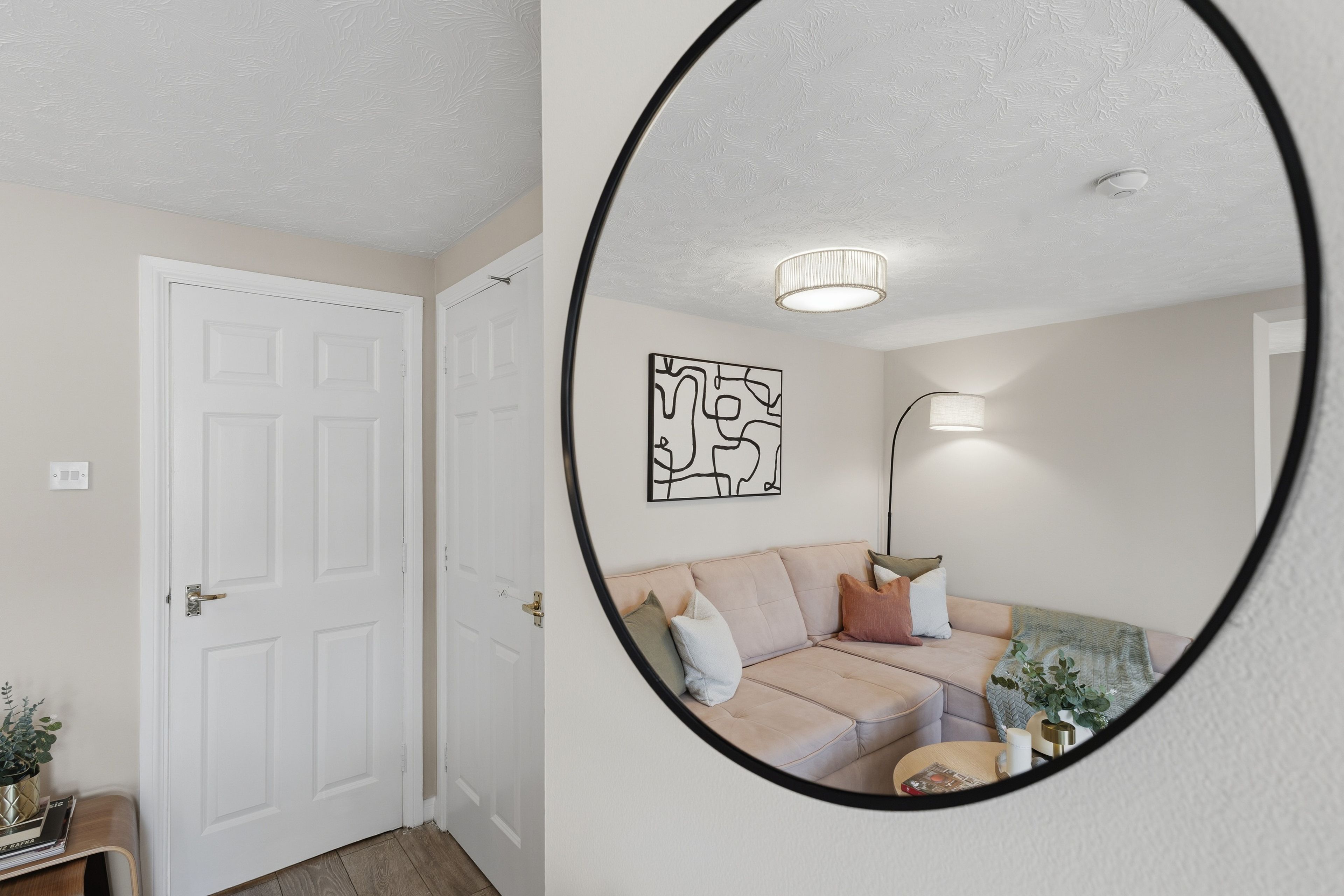 A room with a white door, a sofa with cushions, a wall-mounted abstract art piece, a floor lamp, and a mirror reflecting the room.