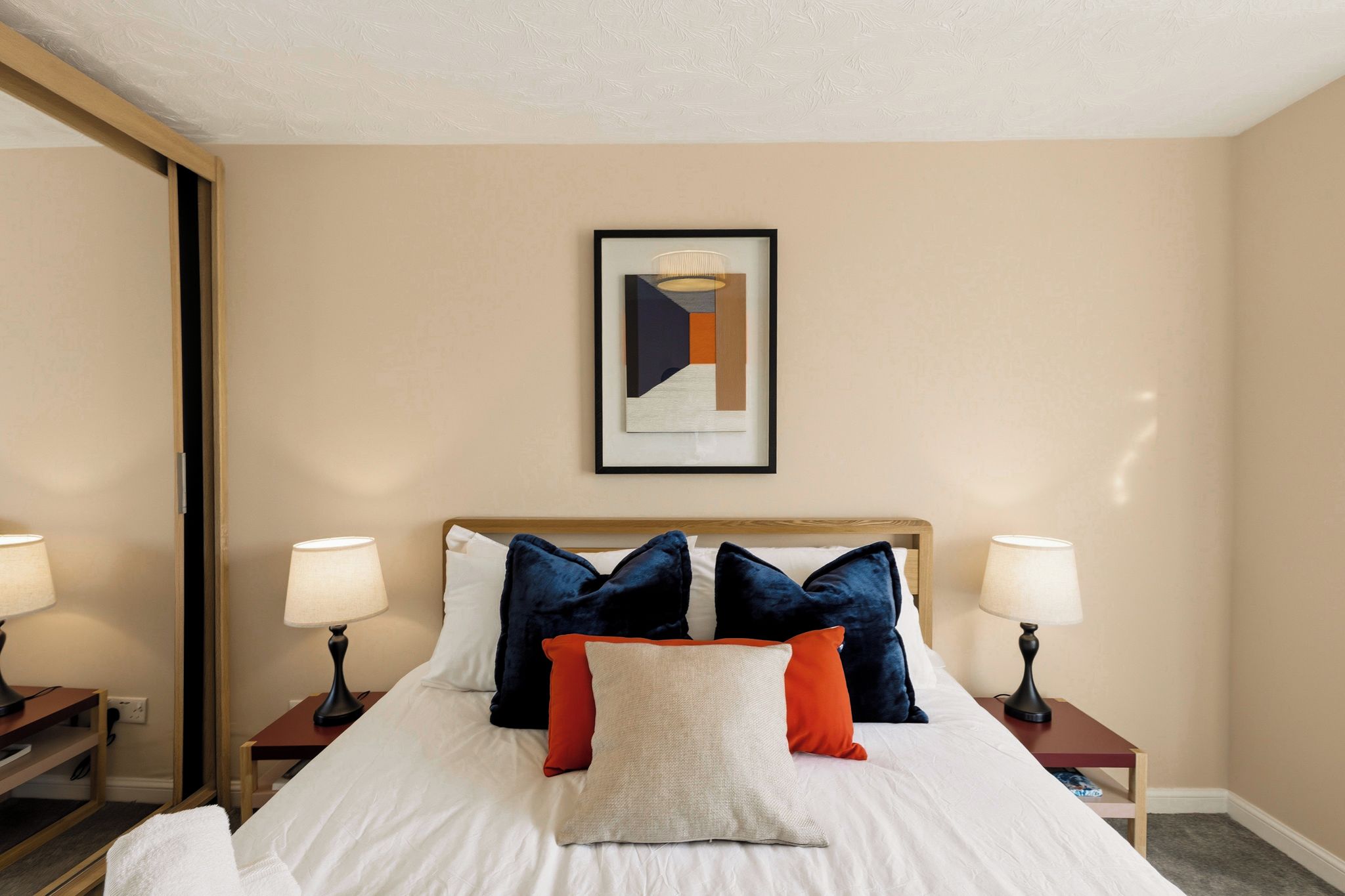 A hotel room with a large bed, two bedside tables with lamps, a framed abstract artwork on the wall, and a mirror on one wall.