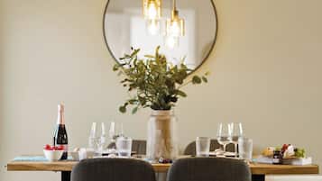 A dining area with a round mirror, two chairs, a table set with glasses, a bottle, and a vase with greenery.