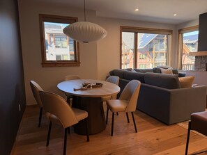 A modern living room with a round dining table, beige chairs, a grey sofa, and a large window providing a view of buildings. - New | Modern Mountain Retreat | Walk to Town (Canmore)