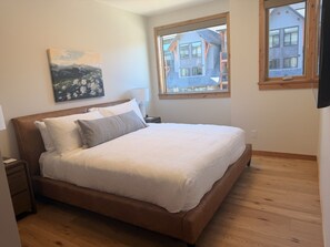 A bedroom with a large bed, a nightstand, a wall-mounted painting, and a window with a view of buildings. - New | Modern Mountain Retreat | Walk to Town (Canmore)