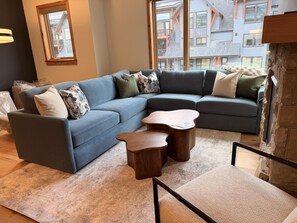 A modern living room with a sectional sofa, two wooden side tables, and a view of a building through a window. - New | Modern Mountain Retreat | Walk to Town (Canmore)